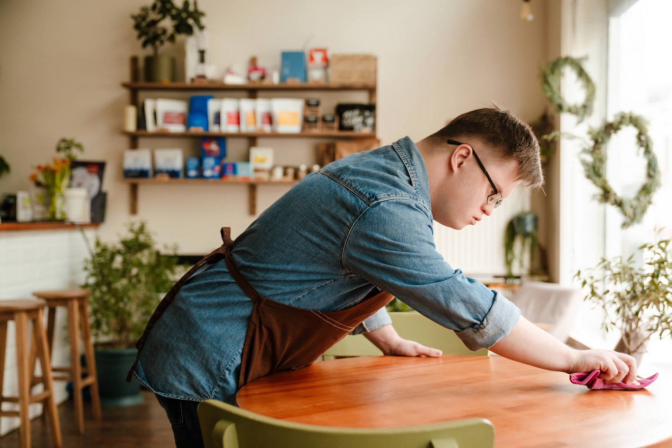 Ein junger Mensch mit Down-Syndrom wischt einen Tisch in einem Café ab. Er trägt ein Hemd und eine Schürze. Im Hintergrund sind Pflanzen und Bücherregale sichtbar, die eine gemütliche Atmosphäre schaffen.