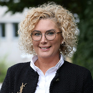 Eine lächelnde Frau mit lockigem, blondem Haar trägt eine schwarze Jacke über einem weißen Oberteil. Sie hat eine Brille auf und steht draußen vor einem unscharfen Hintergrund. Ihr Ausdruck ist freundlich und selbstbewusst.