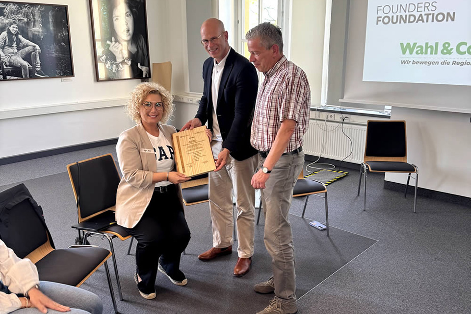 Eine Person mit lockigem Haar überreicht in einem Raum mit Stühlen ein Dokument an zwei Männer. Der Raum hat große Fenster und Fotos an der Wand. Auf dem Bildschirm im Hintergrund steht der Text „Ein großes Dankeschön“ und Logos von Organisationen.